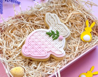 May include: A pink and white bunny-shaped cookie decorated with a white lace pattern and a green flower. The cookie is sitting in a basket of shredded paper.