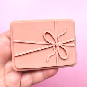 May include: A light pink silicone mould shaped like a gift box with a bow on top. The mould is designed for making polymer clay or resin crafts.