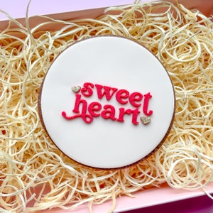 May include: A white round cookie with pink icing and the words "sweetheart" in pink lettering. There are two small gold hearts on the cookie.