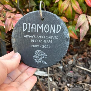 May include: A round, black slate garden marker with the name "DIAMOND" in white script. The text "ALWAYS AND FOREVER IN OUR HEART 2009-2024" is also inscribed. A decorative snake design is at the bottom. A metal stake is attached.