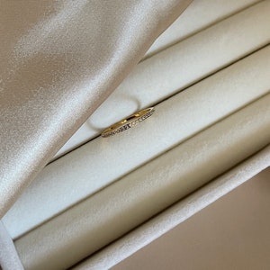 Könnte beinhalten: Ein goldfarbener Ring mit einem Band aus kleinen, klaren Steinen. Der Ring liegt auf einer weißen, samtartigen Oberfläche und einem beigefarbenen Satinstoff.