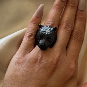 Puede incluir: Anillo negro con un diseño detallado de cabeza de tigre. El anillo, que se lleva en un dedo, muestra los intrincados detalles tallados de la cara del tigre, incluyendo los ojos, la nariz y la textura del pelaje. El anillo es de un material oscuro.