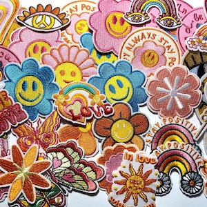 May include: A collection of embroidered fabric patches featuring colorful designs including smiley faces, flowers, rainbows, butterflies, hearts, and peace signs. The patches display various text such as "PEACE LOVE," "STAY POSITIVE," and "in love."