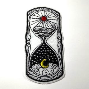 May include: An embroidered patch shaped like an hourglass with black thread outlines on a white background. The top bulb features a red sun with radiating lines above stylized waves. The bottom bulb contains a yellow crescent moon over clouds.
