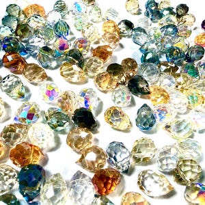 May include: A collection of faceted crystal beads in various colors, including clear, amber, blue, and iridescent. The beads are scattered, showcasing their multiple facets and light refraction. These beads are suitable for jewelry making and crafts.
