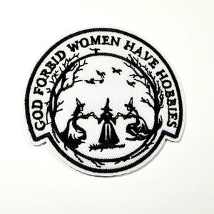 May include: A circular, white patch with black embroidered text and imagery. The text reads "GOD FORBID WOMEN HAVE HOBBIES" around the edge. The center features three figures in witch hats holding hands under a tree.