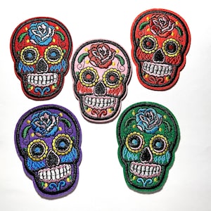 May include: Five embroidered patches in the shape of sugar skulls. Each skull features intricate floral designs and vibrant colors, including red, pink, purple, and green. The patches are detailed with various colors and patterns.