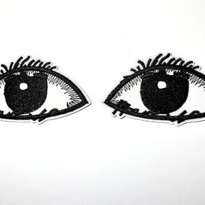 May include: Two embroidered eye patches. Each patch features a black pupil, surrounded by a white iris and black eyelashes. The patches are on a white background.