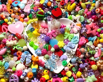 May include: A large assortment of colorful plastic beads and charms, including hearts, stars, flowers, bows, and fruit shapes. The beads are arranged in a white bowl and scattered around it.