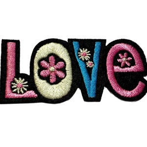 May include: Embroidered patch spelling "LOVE" in a whimsical font. The letters are outlined in black with pink, light blue, and cream-colored floral accents. The patch is designed to be ironed on or sewn onto fabric.