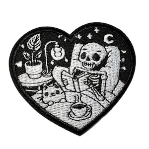 May include: Heart-shaped patch with a black border featuring a skeleton in bed reading a book. A small table with a plant, lamp, and cat is beside the bed. A cup of coffee sits on a saucer. The background is a starry night.