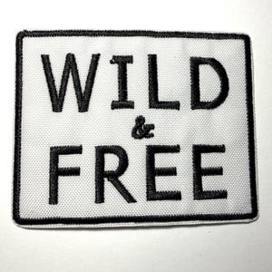 May include: A rectangular patch with the words "WILD & FREE" embroidered in black on a white background. The patch has a black border and is likely designed to be sewn or ironed onto clothing or accessories.