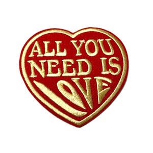 May include: A heart-shaped embroidered patch with a red background and a gold border. The text ALL YOU NEED IS LOVE is stitched in gold thread across the center of the heart, with the word LOVE positioned at the bottom in a larger font size.