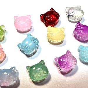 May include: A collection of translucent cat-shaped beads in various colors, including pink, red, yellow, blue, green, and purple. The beads are arranged on a white surface, showcasing their glossy finish and detailed facial features.