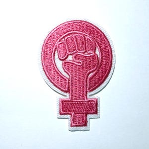 May include: Embroidered patch featuring a pink fist inside a circle, atop a cross-shaped symbol, all outlined in white. The design is a symbol of female empowerment and activism, suitable for clothing or accessories.