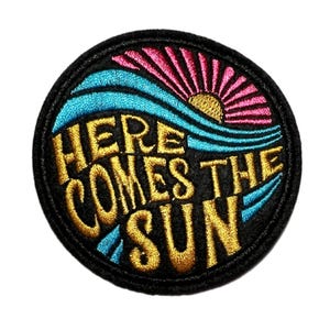May include: Embroidered patch with the phrase "HERE COMES THE SUN" in gold lettering. The design features a sun, waves, and a pink and blue color scheme, all set against a black background. The patch is circular.