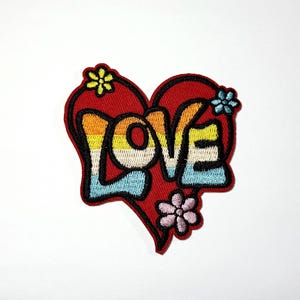 May include: A heart-shaped embroidered patch with a red background and a black border. The word LOVE is stitched in the center with horizontal stripes of orange, yellow, white, and light blue. Three small flowers in yellow, blue, and pink are attached.