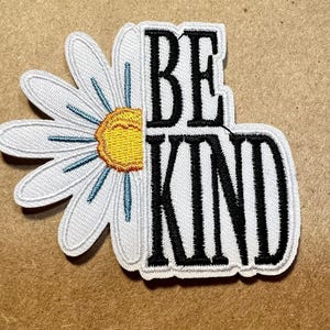 May include: Embroidered patch featuring a white daisy with a yellow center and blue accents, combined with the words "BE KIND" in black, outlined in white. The patch is designed for application on clothing or accessories.