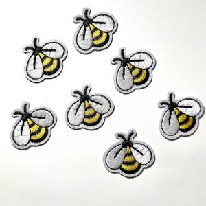 May include: Embroidered bee patches with white, yellow, and black details. These small, decorative patches feature a bee design with a white outline, yellow body, and black accents. Perfect for adding to clothing or accessories.