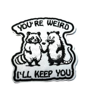 May include: Embroidered patch featuring a raccoon and opossum holding hands. The patch has the text "YOU'RE WEIRD I'LL KEEP YOU" in black lettering on a white background. The patch has a black border.