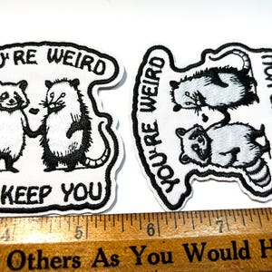 May include: Two embroidered patches with the text "YOU'RE WEIRD I'LL KEEP YOU". One patch features a raccoon and opossum holding hands. The other patch has two raccoons and an opossum. The patches are white with black stitching.
