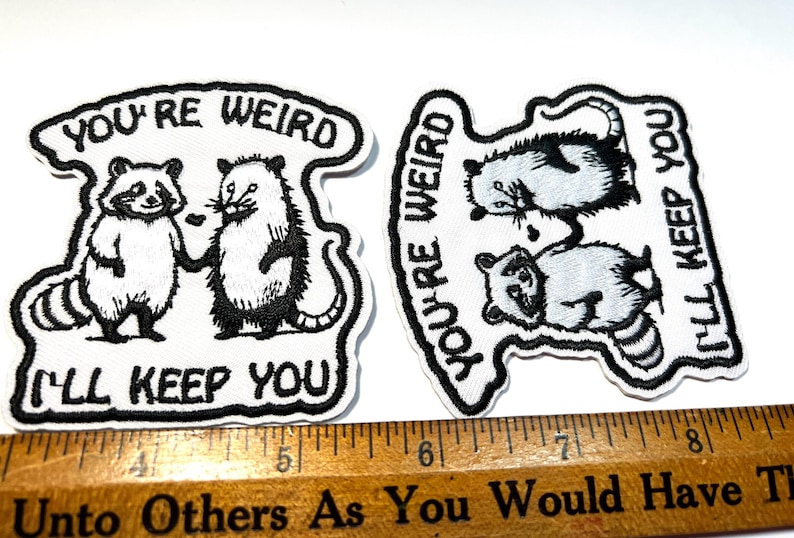 May include: Two embroidered patches with the text "YOU'RE WEIRD I'LL KEEP YOU". One patch features a raccoon and opossum holding hands. The other patch has two raccoons and an opossum. The patches are white with black stitching.