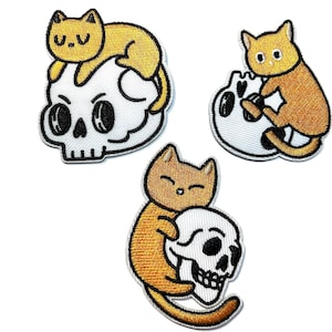 May include: Three embroidered patches featuring orange cats interacting with white skulls. The cat patches have black outlines and are on a white background. The skull patches have black eyes and teeth.