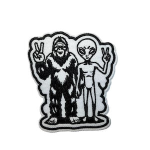 May include: Embroidered patch featuring a black and white illustration of a Bigfoot and an alien, both making peace signs. The patch has a white border and is suitable for jackets, bags, or other fabric items.