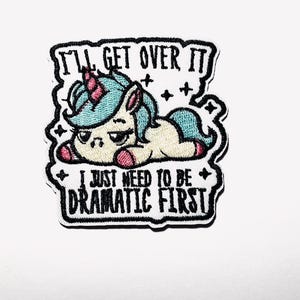 May include: Embroidered patch featuring a cartoon unicorn with a turquoise mane and tail, a pink horn and hooves, and the text "I'll get over it, I just need to be dramatic first." The patch has a black border and is on a white background.