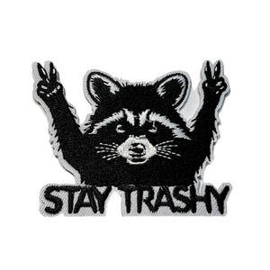 May include: Embroidered patch featuring a black and white raccoon with its arms raised in a peace sign gesture. The text "STAY TRASHY" is displayed in white letters below the raccoon. The patch has a gray border.