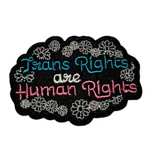 May include: Embroidered patch with the text "Trans Rights are Human Rights" in blue and pink on a black background. The patch has a decorative silver border with floral accents. A statement of support.