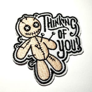 May include: Embroidered patch featuring a voodoo doll design in tan and black, with a stitched appearance. The patch includes the text "Thinking of You!" in a stylized font, outlined in black.