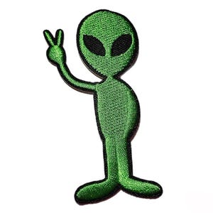 May include: A green embroidered alien patch with black eyes and a peace sign gesture. The alien is standing upright with a black outline, and the patch is on a white background. This patch is perfect for jackets, backpacks, or other fabric items.
