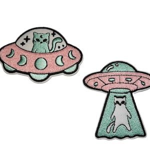 May include: Two embroidered patches featuring a cat theme. One shows a cat in a pink and mint green UFO, the other depicts a cat being beamed up by a UFO. The patches have a black border.