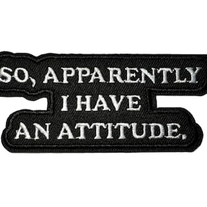 May include: Black embroidered patch with the text "SO, APPARENTLY I HAVE AN ATTITUDE." The patch has a black background with white stitching. The text is in a bold, sans-serif font.