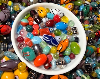 May include: A white ceramic bowl filled with a large assortment of multi-colored glass beads in various shapes and sizes. The collection includes round, oval, and cylindrical beads in shades of red, blue, green, orange, yellow, and black.