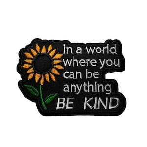 May include: Embroidered patch with a black background, featuring a sunflower and the text "In a world where you can be anything BE KIND" in white. The sunflower has yellow petals and a dark center.