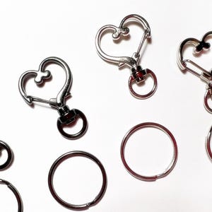 May include: Five silver and black heart-shaped keychains with a swivel clasp and a ring. The keychains are on a white background.