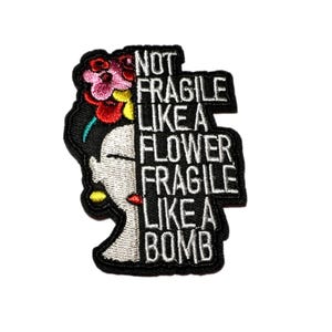 May include: Embroidered patch featuring a stylized portrait of a woman with a floral headdress. The patch is black with white text that reads "NOT FRAGILE LIKE A FLOWER FRAGILE LIKE A BOMB."