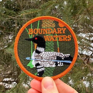 May include: A round embroidered patch featuring a loon in a lake scene. The patch has an orange border and the words "BOUNDARY WATERS" in gold. The loon has black, white, and gray feathers, with a red eye.