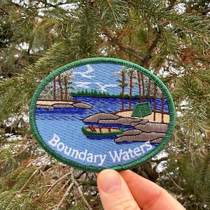 May include: An embroidered patch with a scenic landscape design. The patch features a blue lake, trees, and rocks, with the text "Boundary Waters" in white. The patch has a green border and is held against a backdrop of evergreen branches.