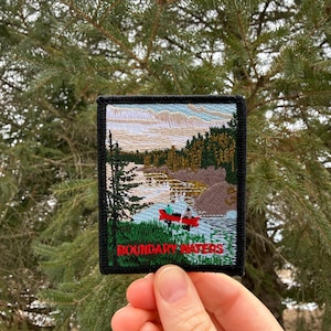May include: Embroidered patch featuring a scenic landscape with a lake, trees, and a red canoe with two people. The patch has a black border and the words "BOUNDARY WATERS" in red and green.