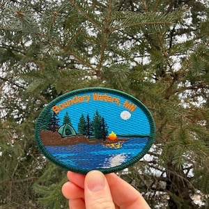 May include: An oval patch with the text "Boundary Waters, MN" in orange lettering. The patch features a camping scene with a tent, trees, a campfire, and a moon over a blue lake. The patch is surrounded by a green border.