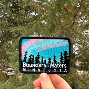 May include: Embroidered patch featuring a landscape scene with a teal and pink sky above a silhouette of trees. The text "Boundary Waters Minnesota" is displayed in white. The patch has a black border.
