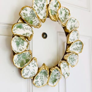Peut inclure: Une couronne décorative composée de coquilles d'huîtres. Les bords des coquilles sont dorés et présentent un motif floral vert sur fond blanc. La couronne est fixée sur une base circulaire marron.