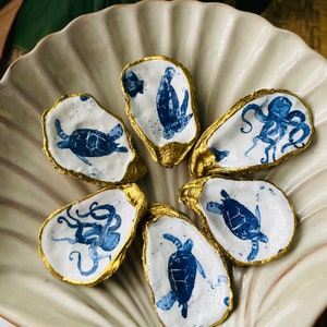 May include: Six white oyster shells with gold rims, each painted with a blue sea creature. The shells are arranged in a circle on a white, scalloped dish.