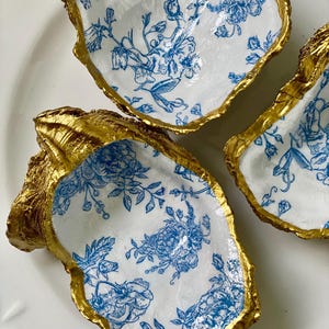 May include: Three oyster shell dishes with a gold-painted rim and a blue floral pattern on a white background. The shells are arranged on a white surface, showcasing the unique shape and design of each piece.