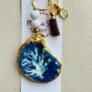 May include: A handmade keychain featuring a painted oyster shell with a blue and white coral design, accented with gold trim. The keychain includes a gold-tone clasp, a brown tassel, and white wooden beads. The product is attached to a white card.