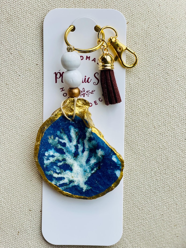 May include: A handmade keychain featuring a painted oyster shell with a blue and white coral design, accented with gold trim. The keychain includes a gold-tone clasp, a brown tassel, and white wooden beads. The product is attached to a white card.