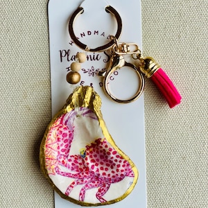 May include: Handmade oyster shell keychain with a gold-painted edge and a pink crab design. The keychain has a gold-colored ring, a pink tassel, and wooden beads. The background is a white card with text.